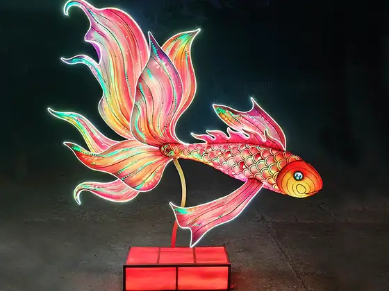Carpa luminosa LED de cola larga y velos amplios, iluminada en rosas, naranjas y verdes sobre una peana roja