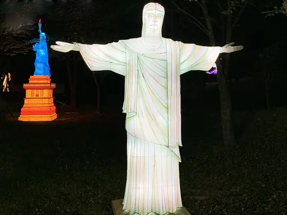 Réplica luminosa LED del Cristo Redentor con los brazos abiertos, iluminada en blanco entre los árboles de un parque de noche