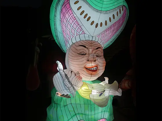 Figura luminosa LED de la duquesa con un enorme tocado verde y rosa, vestido verde y un gato en brazos, iluminada desde dentro